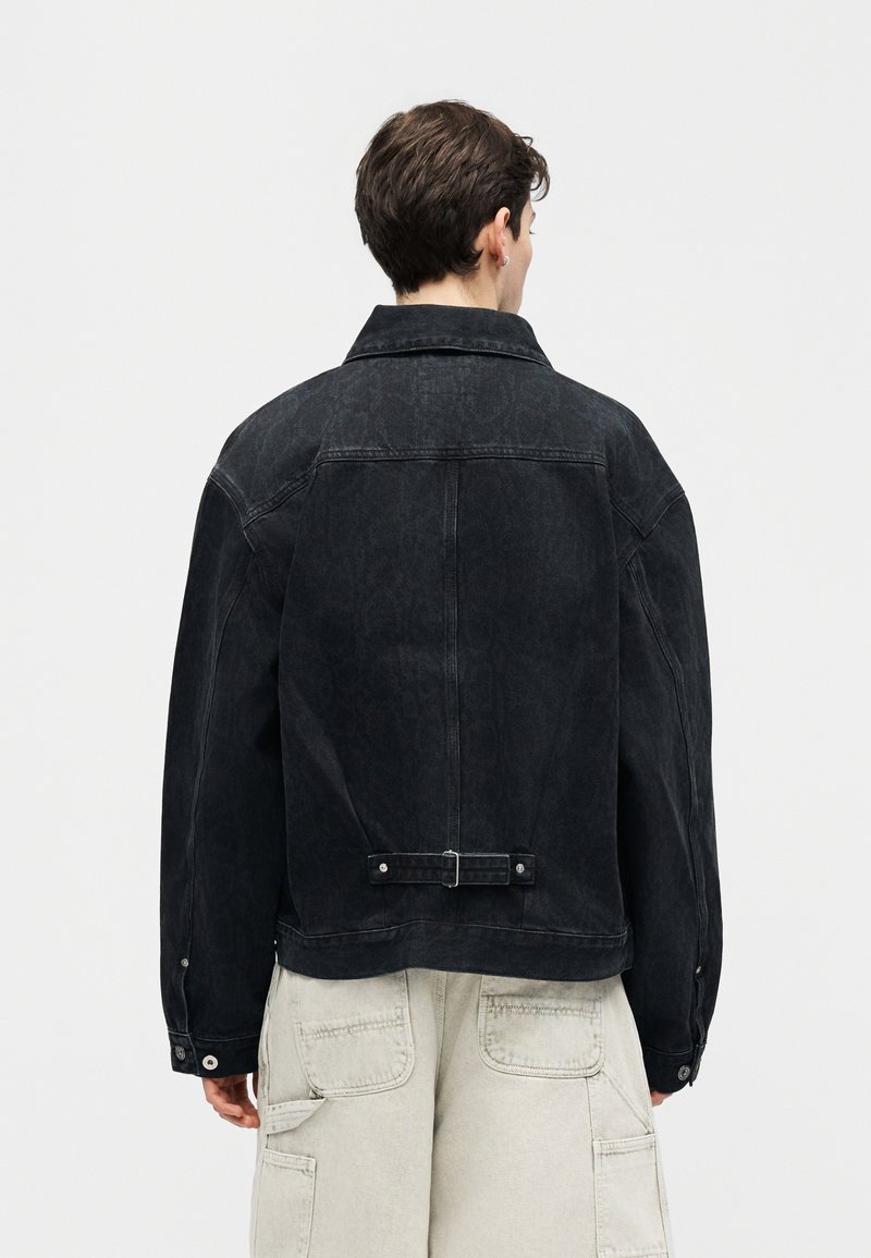 Persoon met kort donker haar, gekleed in een zwarte spijkerjack en lichtbeige cargobroek, gezien van achteren tegen een effen achtergrond.