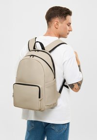 Beige hátizsák fekete kiegészítőkkel, sima felülettel, két felső cipzárral, elülső zsebbel és állítható pántokkal. Egy személy hátán viselve.