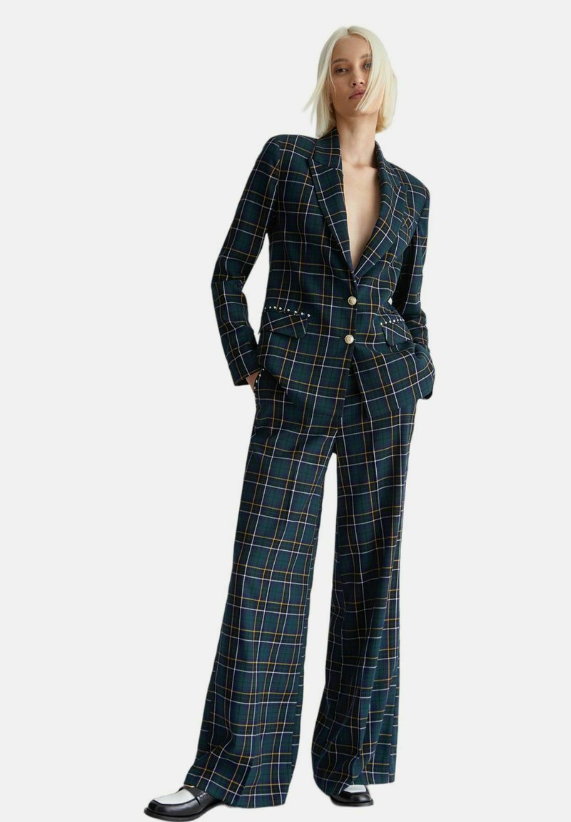 Completo giacca e pantaloni a quadri verde scuro con gamba larga, caratterizzato da strisce arancioni e gialle e dettagli in bottoni metallici. Finitura in tessuto testurizzato.