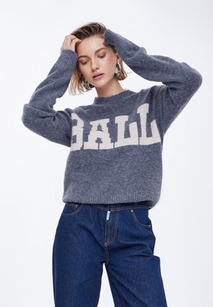 Grå stickad tröja med krämfärgad "BALL" text, rund hals, långa ärmar och en mjuk struktur, i kombination med mörkblå denimjeans.