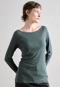 Ilga rankovė viršūnė muted green spalvos, su plačiu laivapriekio kaklu, minkšta tekstūra ir prigludusia siluetu. Lygi siūlė ir subtilus blizgesys.