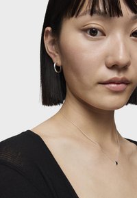 Orecchini a cerchio in argento e una collana delicata con un piccolo pendente nero indossati con un top nero texturizzato. Inquadratura ravvicinata del collo e del viso della modella.