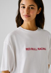 Ung kvinde iført en hvid T-shirt med teksten "RED BULL RACING" i rødt, der står foran en ensfarvet lys grå baggrund.