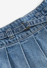 Nahaufnahme von blauem Denimstoff mit eingenähten Falten und einer Gürtelschlaufe, die einen schmalen Gürtel hält, der mit einem doppelten Metallring befestigt ist.