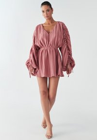 Ein Modell steht in einem lockeren, pinken Kleid mit langen Ärmeln und tailliertem Schnitt, das einen eleganten Stil zur Schau stellt.