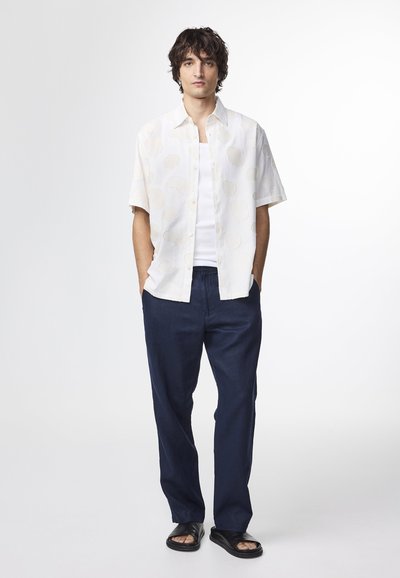 Witte kortärmige shirt met een reliëf schelpenpatroon, gedragen over een witte tanktop; gecombineerd met marineblauwe linnenbroek en zwarte sandalen.