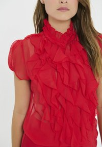 Czerwona przezroczysta bluzka z wysokim, falbaniastym kołnierzem oraz warstwowym detalem falbanek z przodu. Krótkie rękawy i lekki materiał.