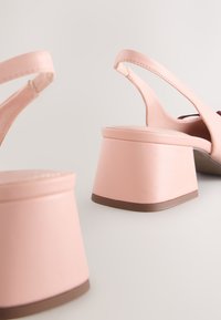 Par de sandalias slingback de tacón ancho color rosa claro con acabado liso y bordes marrones en la suela sobre una superficie clara.