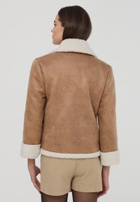 Giacca in finta pelliccia shearling marrone con colletto e polsini bianchi, caratterizzata da una texture liscia e una vestibilità rilassata. L'orlo della giacca termina in vita.
