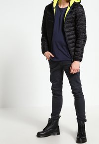 Person in schwarzer Daunenjacke mit neongrünem Kapuzen, dunkelblauem Hemd, schwarzen Skinny-Jeans und schwarzen Schnürstiefeln, der vor einem weißen Hintergrund steht.