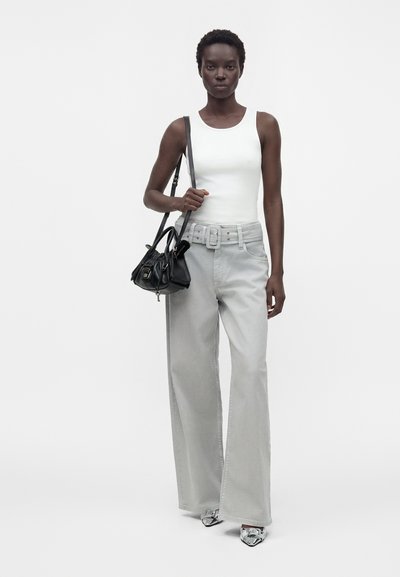 Femme portant un haut blanc sans manches, un pantalon gris clair taille haute à jambes larges avec ceinture, des chaussures argentées pointues, tenant un sac à épaule noir.
