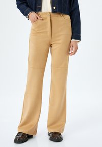 Pantalones acampanados en tono beige con un acabado de tela suave, cintura alta y bolsillos laterales. Combinados con zapatos oscuros y gruesos para crear contraste.
