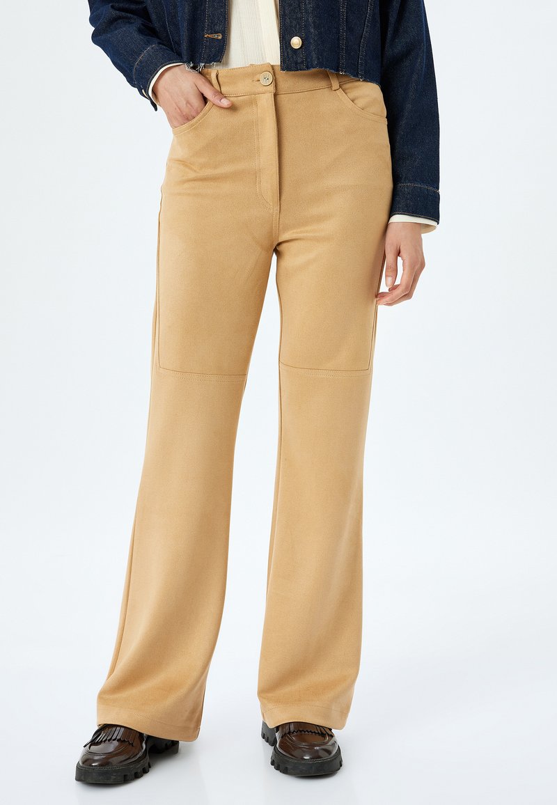 Pantalones acampanados en tono beige con un acabado de tela suave, cintura alta y bolsillos laterales. Combinados con zapatos oscuros y gruesos para crear contraste.
