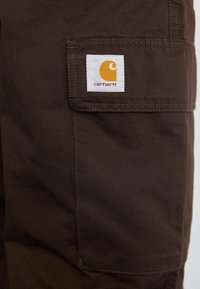 Brązowe spodnie cargo wykonane z trwałego materiału, z widoczną kieszenią naszywaną z białą metką i pomarańczowym logo, podkreślające praktyczny design.