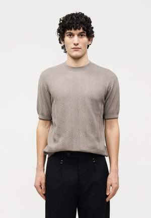 Άνδρας με σγουρά μαλλιά, φορώντας ένα μπεζ κοντομάνικο πλεκτό πουλόβερ με υφή και μαύρο παντελόνι, στέκεται μπροστά σε ένα απλό ανοιχτό φόντο.