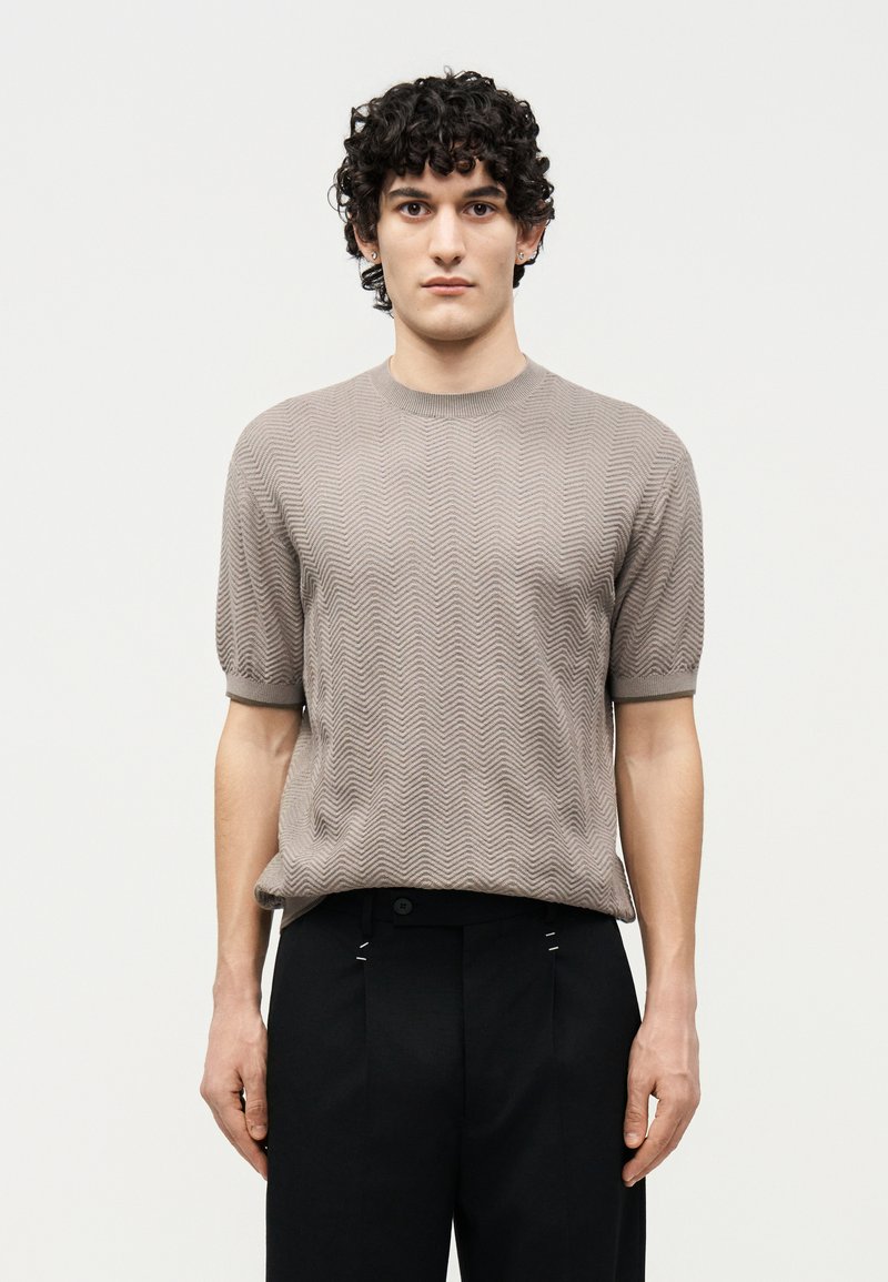 Άνδρας με σγουρά μαλλιά, φορώντας ένα μπεζ κοντομάνικο πλεκτό πουλόβερ με υφή και μαύρο παντελόνι, στέκεται μπροστά σε ένα απλό ανοιχτό φόντο.