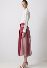 Femme en blouse blanche à manches longues et jupe rose à mi-mollet avec superposition rouge transparente, se tenant de profil, portant des sandales à talons hauts blanches.