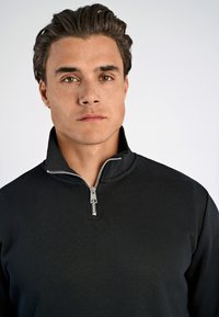 Maglione zip-up nero con colletto alto e dettagli in cerniera argentati. Texture liscia e design aderente, caratterizzato da un colore uniforme.