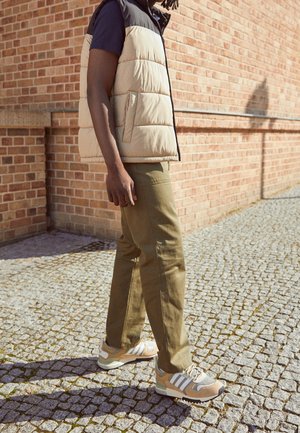 Person wearing beige puffer vest, navy shirt, olive pants, and beige Adidas sneakers walking on a cobblestone path by a brick wall.