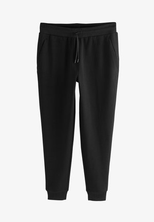 Homme portant un t-shirt noir, un pantalon de jogging noir et des baskets noires, debout sur un fond gris.