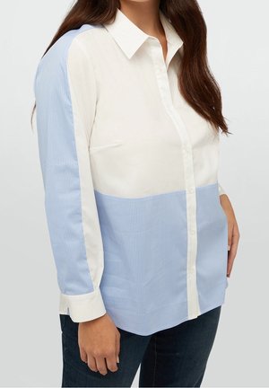 Woman wearing a button-up shirt with white upper and striped light blue lower sections and sleeves, paired with dark pants.