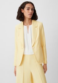 Yellow blazer with a single button closure, notch lapels, and front pockets, paired with a white top featuring a keyhole neckline.