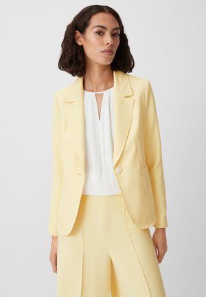 Yellow blazer with a single button closure, notch lapels, and front pockets, paired with a white top featuring a keyhole neckline.