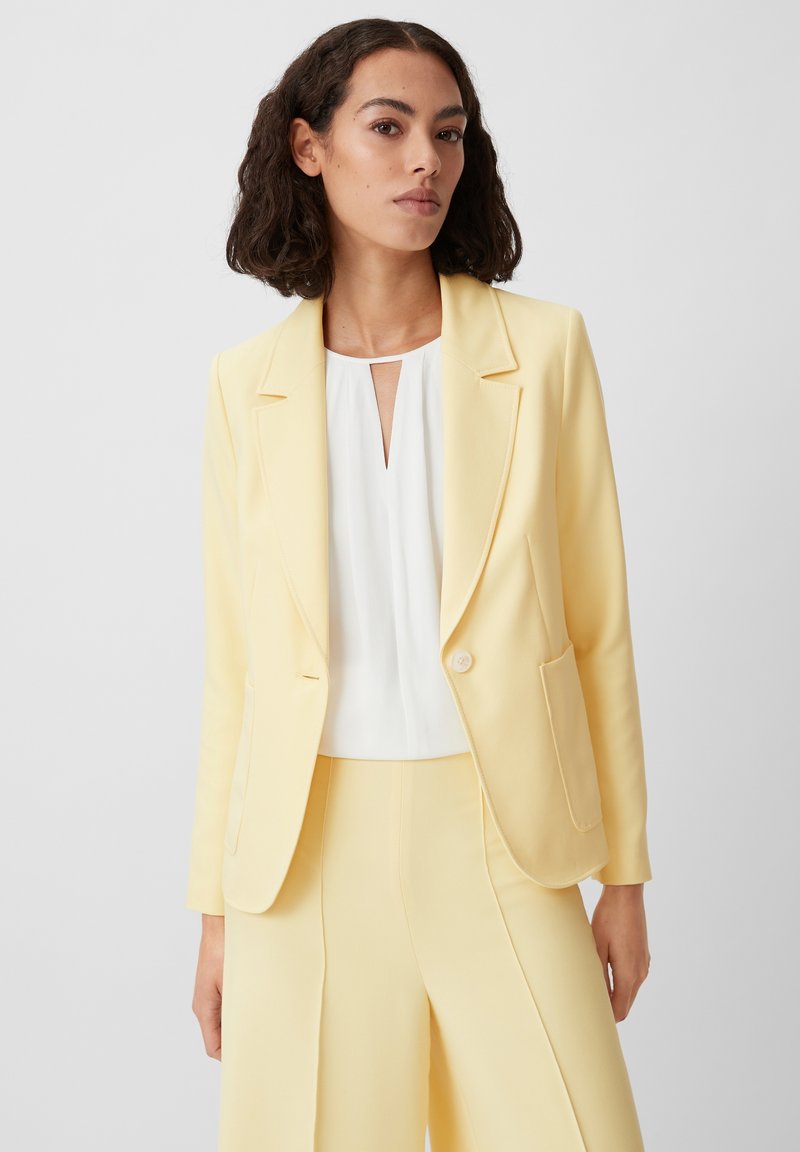 Yellow blazer with a single button closure, notch lapels, and front pockets, paired with a white top featuring a keyhole neckline.