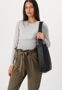 Sac à épaule en cuir noir avec une texture lisse, une large bandoulière et un design minimaliste. Le modèle porte un haut moulant gris à manches longues et un pantalon olive.