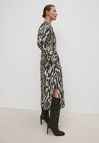 Langes schwarzes und cremefarbenes Kleid mit einem wellenförmigen Muster, langen Ärmeln und seitlichen Schlitzen, getragen mit knielangen schwarzen Stiefeletten mit Absatz, glatte Stofftextur.