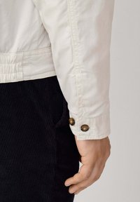 Primer plano de la mano de una persona cerca de la muñeca de una chaqueta color crema con dos botones de metal, combinada con pantalones negros de canalé.