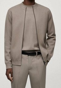 Light beige zip-up sweatshirt with a round neckline, textured fabric, and a slim fit, paired with matching trousers and a black belt.
