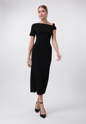 Abito midi nero aderente con design monospalla e dettaglio a nodo sulla spalla; realizzato in tessuto liscio. Stile elegante con tacchi.