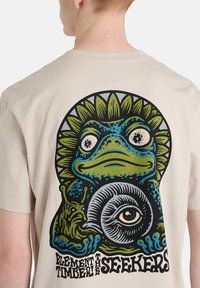 Ανοιχτό μπεζ μπλουζάκι με πολύχρωμο γραφικό βατράχου, που διαθέτει μοτίβο ματιού και φυλλώδη διακοσμητικά. Ο σχεδιασμός έχει έντονες περιγράμματα και περίτεχνες λεπτομέρειες.