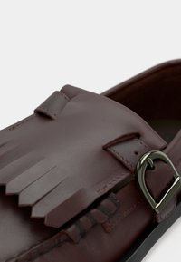 Mocassin en cuir marron avec des détails à franges et une sangle en boucle argentée sur une semelle noire, montré en gros plan.