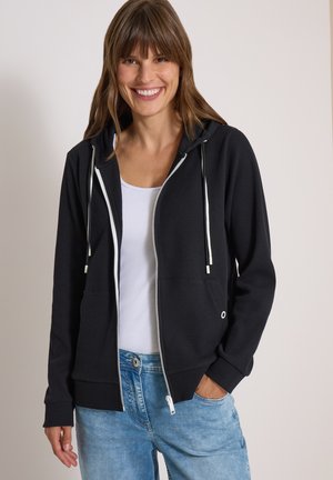 Femme souriante portant un sweat à capuche noir zippé avec des cordons blancs, une chemise blanche et un jean bleu clair, une main dans la poche.