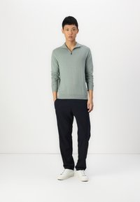 Light green zip neck sweater with long sleeves, paired with dark navy pants and white sneakers, features a fitted design and ribbed cuffs.
