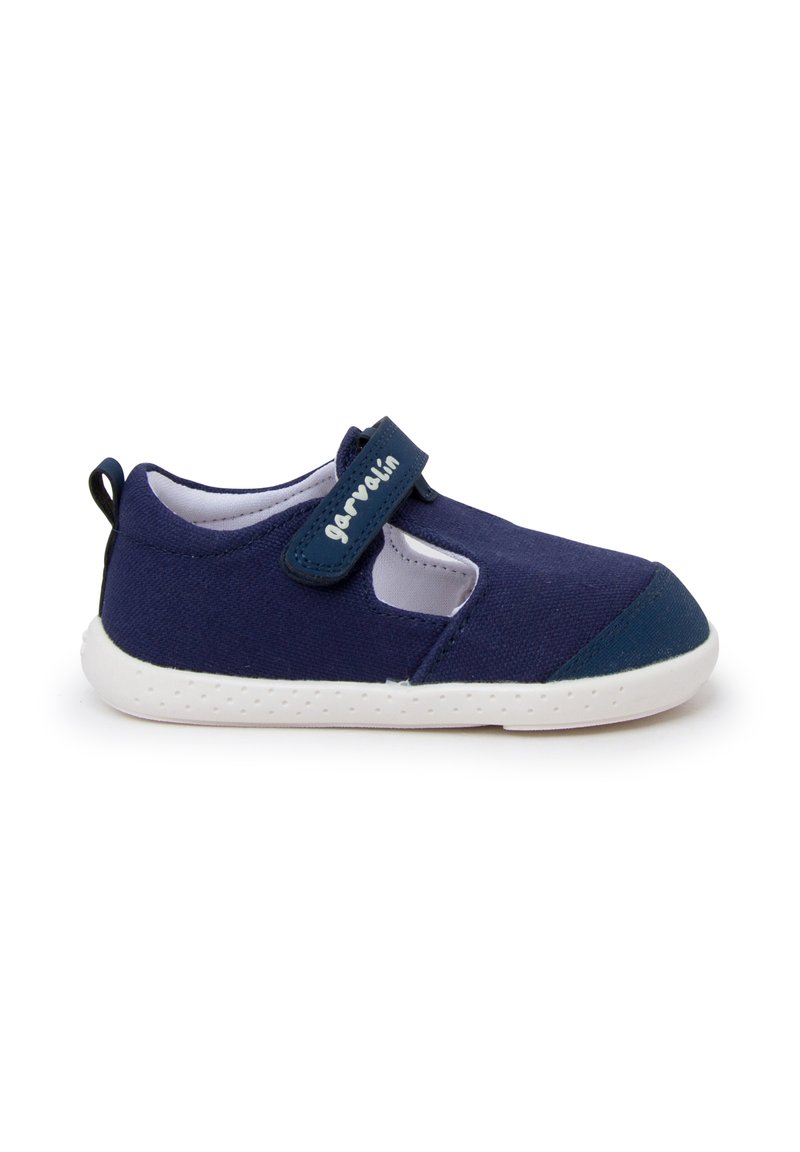 Zapato de lona azul marino con tira de velcro, suela blanca, puntera redondeada, lados cortados y una pequeña lengüeta en el talón para facilitar el uso.