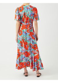 Femme portant une longue robe fleurie avec des fleurs rouges et violettes et des feuilles vertes, se tenant de dos, portant des sandales noires.