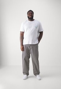 Hombre de pie con una camiseta gráfica blanca, pantalones holgados a rayas y zapatillas blancas frente a un fondo claro y sencillo.