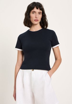 Woman with dark hair wearing a black short-sleeve top with white trim and white high-waisted pants, standing with one hand in pocket.