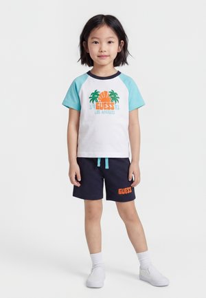 Jeune fille debout face à l'avant, portant un T-shirt blanc et turquoise avec un motif de palmier et un short bleu marine avec le logo "GUESS".