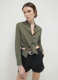 Camicia verde oliva con bottoni e davanti annodato, dotata di maniche lunghe e tessuto testurizzato. Abbinata a pantaloni shorts neri e una cintura intrecciata.