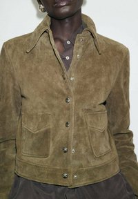 Veste en daim marron avec un col classique, fermeture à boutons et deux poches avant à rabat pointu. Texture douce et coupe ajustée.