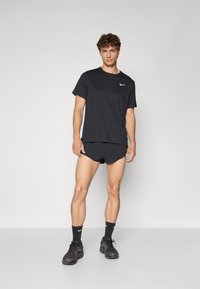 Camiseta negra de Nike con mangas cortas, combinada con pantalones cortos negros. El modelo lleva calcetas y zapatos negros, de pie sobre un fondo gris claro.