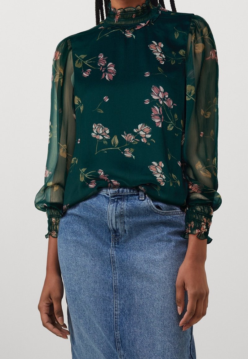 Femme portant un chemisier floral transparent vert foncé avec col et poignets à volants, rentré dans une jupe en jean bleue taille haute.