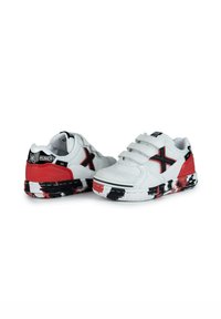 Zapatillas sintéticas blancas con acentos en negro y rojo, con tiras de velcro y una suela texturizada con un patrón de pintura llamativo.