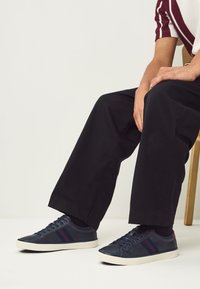 Mørkeblå sneakers med røde striber, glat lædertekstur, hvid gummisål, kombineret med sorte bukser med vide ben. Foden hviler på jorden.