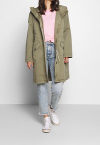 Oliwkowa parka z kapturem, z regulowanym paskiem w talii, dwiema przednimi kieszeniami, nałożona na różową koszulę, zestawiona z jasnoniebieskimi dżinsami w stylu distressed i białymi sneakerami.