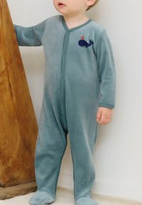 Bambino in tutina blu con design di balena, in piedi e appoggiato a un mobile di legno contro una parete bianca.